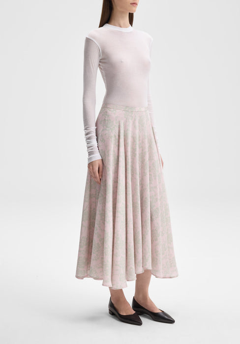 A person wearing a sheer white long-sleeve top layered over a camisole, paired with a mid-length, flowy pink skirt with a delicate floral pattern and black pointed-toe flats.