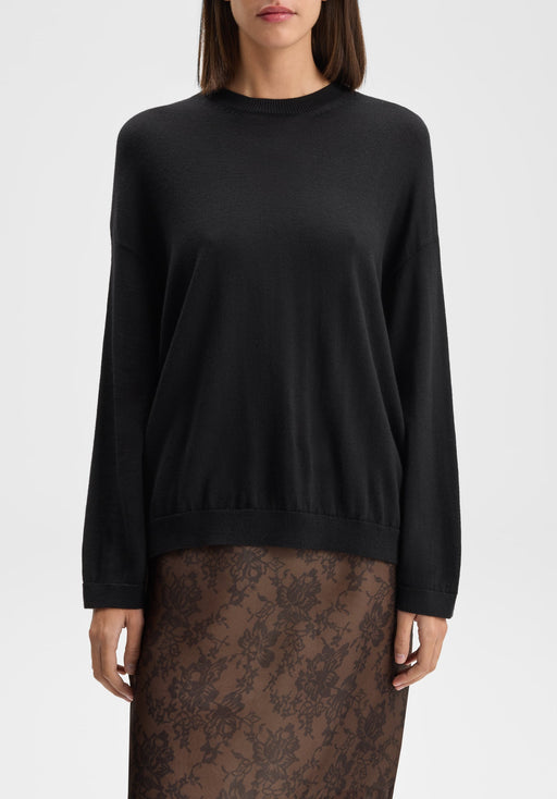 Front-facing mid-length view of a model standing upright with arms relaxed at her sides, wearing a black long-sleeve knit sweater paired with a sheer brown floral lace skirt against a plain background.
