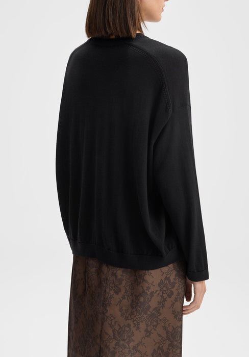 The model is shown from the back in a three-quarter view, with her body angled slightly to the right. Her head is gently turned to the side, and her arms rest naturally by her sides, highlighting the back and drape of the black sweater and the top of the lace skirt.