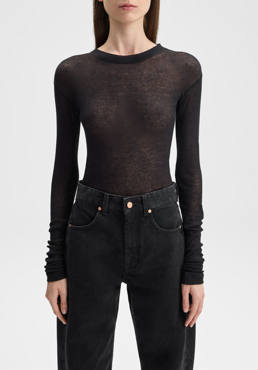 Front-facing close-up of a model from neck to hips, standing upright with arms relaxed at her sides, wearing a sheer black long-sleeve top paired with high-waisted black jeans against a plain background.