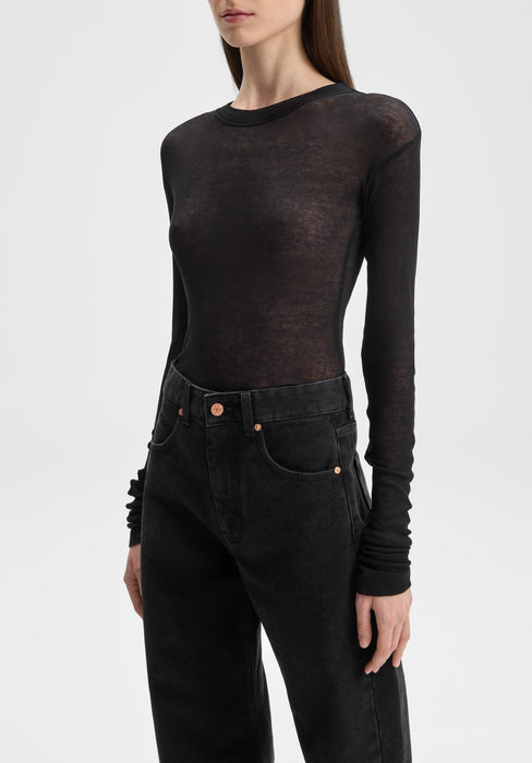 Close-up of a model from shoulders to hips wearing a sheer black long-sleeve top over dark high-waisted jeans, standing slightly angled with one arm relaxed by her side against a plain background.