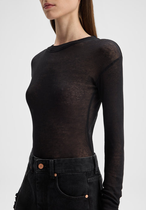 Close-up side view of a model from shoulders to hips, slightly angled to the left, wearing a sheer black long-sleeve top tucked into high-waisted black jeans, with one arm relaxed along her side against a plain background.