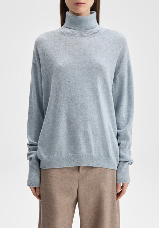 Front-facing mid-length view of a model standing upright with arms relaxed at her sides, wearing a light blue oversized turtleneck sweater paired with beige trousers against a plain background.