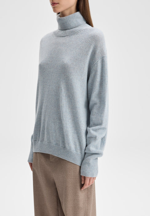 Three-quarter view of the model’s upper body, standing upright with her torso slightly angled to the side, shoulders relaxed, and one arm hanging naturally at her side while the other falls straight down, showcasing the drape of the turtleneck sweater.