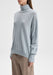 Three-quarter view of the model’s upper body, standing upright with her torso slightly angled to the side, shoulders relaxed, and one arm hanging naturally at her side while the other falls straight down, showcasing the drape of the turtleneck sweater.