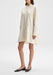 Full-length three-quarter view of a model standing with one leg slightly forward, wearing a cream long-sleeve button-down shirt dress with a flared hem and pointed collar, paired with black flats against a plain background.