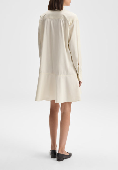 Back-facing full-length view of the model standing upright with her body slightly angled, arms relaxed at her sides, wearing a cream long-sleeve shirt dress with a softly flared hem and black flats against a plain background.