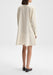 Back-facing full-length view of the model standing upright with her body slightly angled, arms relaxed at her sides, wearing a cream long-sleeve shirt dress with a softly flared hem and black flats against a plain background.