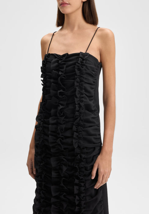 Three-quarter view of the model standing upright, body slightly angled to the side with shoulders turned gently, arms relaxed at her sides, wearing a black spaghetti-strap dress with vertical ruffle detailing.