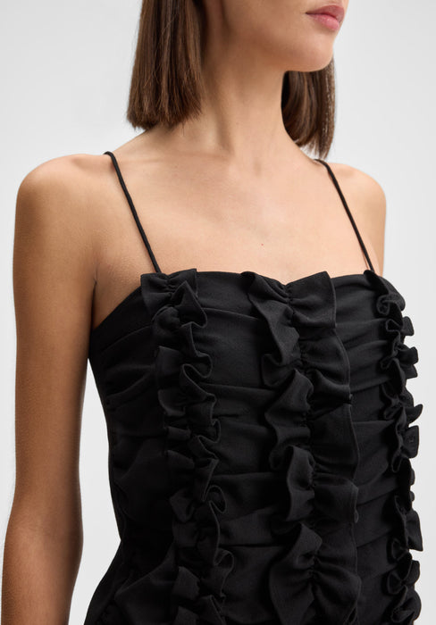 Close-up three-quarter view of the model’s upper body, slightly angled to the side with shoulders turned and head subtly facing forward, wearing a black spaghetti-strap dress with ruffled detailing across the bodice.