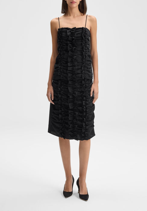 Front-facing full-length view of the model standing upright with feet close together and arms relaxed straight at her sides, wearing a black midi dress with thin straps and vertical ruffle detailing, paired with black heels.