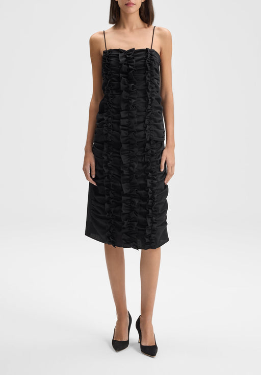 Front-facing full-length view of the model standing upright with feet close together and arms relaxed straight at her sides, wearing a black midi dress with thin straps and vertical ruffle detailing, paired with black heels.