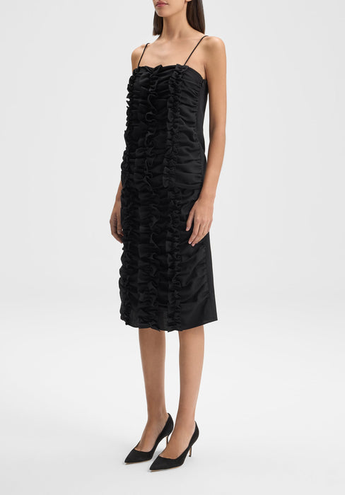 Full-length view of a model standing upright and slightly angled to the side, wearing a black midi dress with thin straps and vertical ruffle detailing down the front, paired with black heels against a plain background.