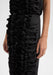 Close-up detail of a black dress showing vertical ruffle embellishments along the fabric, with the model’s hands relaxed at her sides against a plain background.
