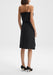 Back view of a model standing upright and slightly angled to the side, wearing a black midi dress with thin straps and a smooth silhouette, paired with black high heels, against a plain background.