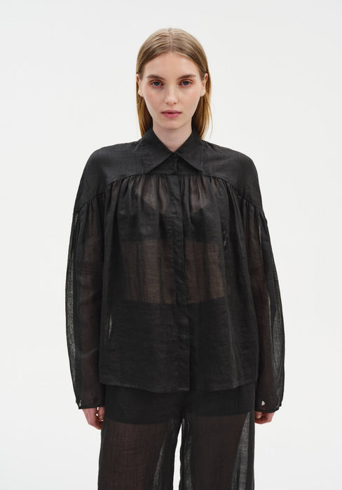 Front view of a model wearing a sheer black long-sleeve blouse with a pointed collar and gathered detailing, paired with matching translucent trousers, emphasizing the lightweight, semi-transparent fabric and relaxed silhouette.