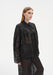 Three-quarter side view of a model wearing a sheer black long-sleeve blouse with a pointed collar and gathered yoke, paired with matching translucent trousers, highlighting the airy fabric and relaxed fit.