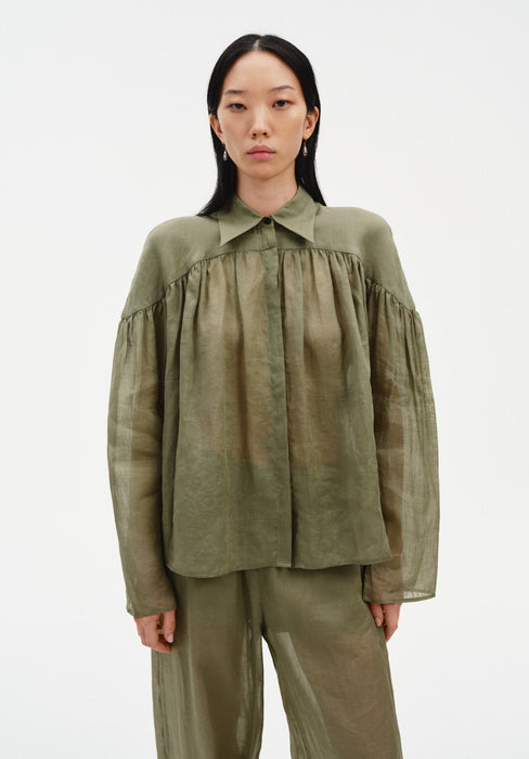 Front-facing view of a model standing upright with arms relaxed at her sides, wearing a sheer olive-green blouse with a pointed collar and gathered detailing, paired with matching lightweight trousers, emphasizing the soft, airy fabric and straight silhouette.