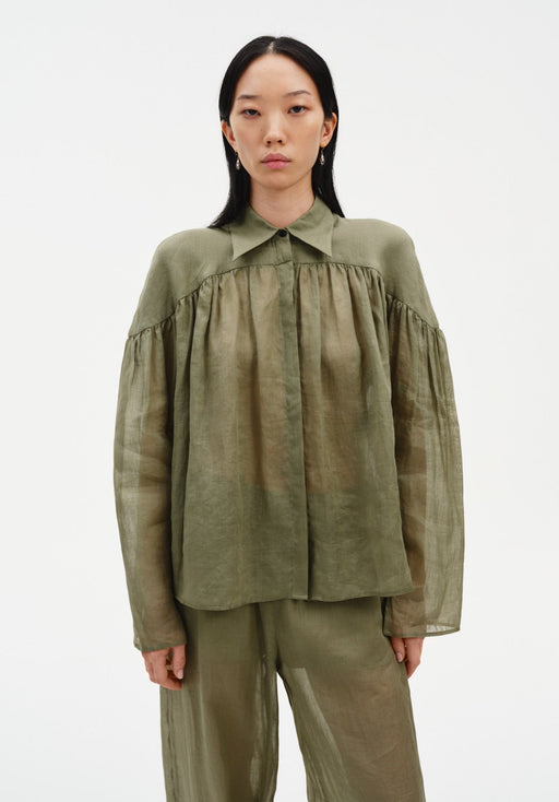 Front-facing view of a model standing upright with arms relaxed at her sides, wearing a sheer olive-green blouse with a pointed collar and gathered detailing, paired with matching lightweight trousers, emphasizing the soft, airy fabric and straight silhouette.