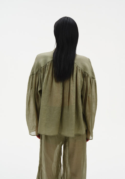 Back-facing view of a model standing upright with arms relaxed at her sides, wearing a sheer olive-green blouse with gathered yoke and long sleeves, paired with matching translucent trousers, showing the loose drape from behind.