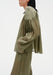 Side view of a model with one hand in her pocket, wearing a sheer olive-green blouse with gathered shoulder detailing and loose, flowing sleeves, paired with matching lightweight trousers, emphasizing the airy fabric and relaxed silhouette.