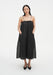 A woman stands facing forward with a relaxed posture against a plain white background, wearing a black sleeveless midi dress with thin straps and a loose, gathered silhouette. Her hands are in her pockets, her dark shoulder-length hair is worn down, and she tilts her head slightly with a neutral expression, paired with black pointed heels.