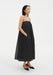 A woman stands in a relaxed three-quarter side pose against a plain white background, wearing a black sleeveless midi dress with thin straps and a loose, gathered silhouette. She has one hand in her pocket, her dark shoulder-length hair worn down, and she looks slightly to the side with a neutral expression, paired with black pointed heels.