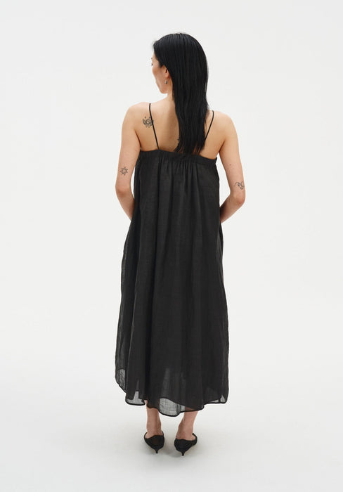 Rear view of a woman standing against a plain white background, wearing a black sleeveless midi dress with thin straps and a gathered, flowing silhouette. Her dark shoulder-length hair falls down her back, and small tattoos are visible on her upper back and arms.