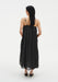 Rear view of a woman standing against a plain white background, wearing a black sleeveless midi dress with thin straps and a gathered, flowing silhouette. Her dark shoulder-length hair falls down her back, and small tattoos are visible on her upper back and arms.
