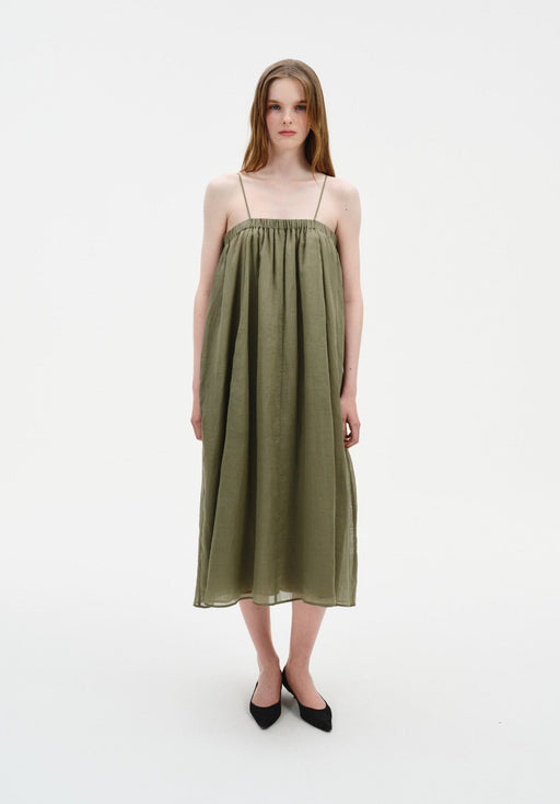 A woman stands facing forward in a straight, centered stance with feet close together, wearing an olive-green sleeveless midi dress with thin straps and a loose, gathered silhouette, paired with black pointed heels.