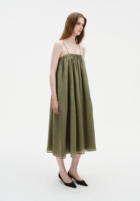 A woman stands in a relaxed three-quarter side pose with a slight forward lean, wearing an olive-green sleeveless midi dress with thin straps and a loose, flowing silhouette, paired with black pointed heels.