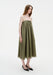 A woman stands in a relaxed three-quarter side pose with a slight forward lean, wearing an olive-green sleeveless midi dress with thin straps and a loose, flowing silhouette, paired with black pointed heels.