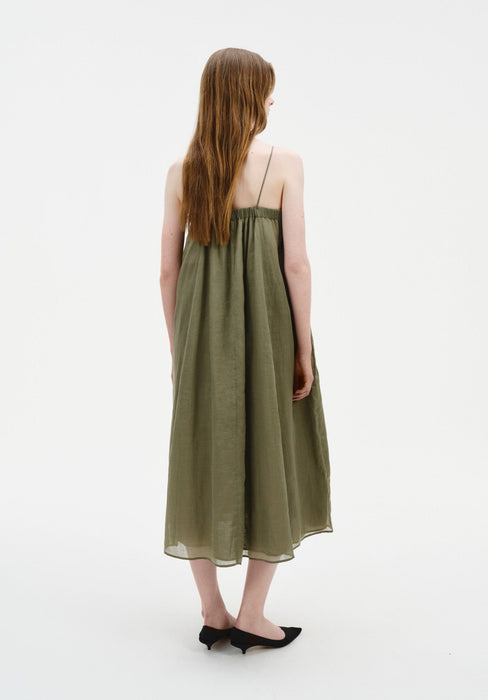Rear view of a woman standing with a slight turn to one side, her weight subtly shifted, wearing an olive-green sleeveless midi dress with thin straps and a loose, gathered silhouette.