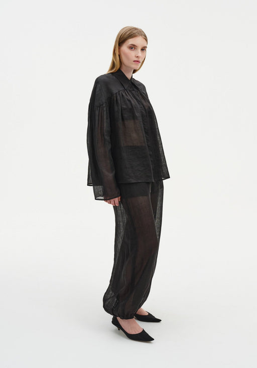 A woman stands in a slight three-quarter side pose against a plain white background, wearing a sheer black loose blouse with gathered shoulders and matching semi-transparent billowy trousers, paired with black pointed heels. Her straight light brown hair is worn down, and she looks toward the camera with a neutral expression.