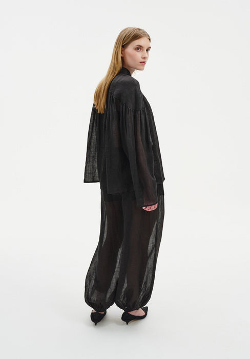 Rear three-quarter view of a woman turning her head slightly toward the camera, wearing a sheer black loose blouse and matching billowy trousers with elasticated cuffs, paired with black heels. Her straight light brown hair falls down her back, and the semi-transparent fabric reveals the silhouette beneath.