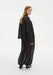 Rear three-quarter view of a woman turning her head slightly toward the camera, wearing a sheer black loose blouse and matching billowy trousers with elasticated cuffs, paired with black heels. Her straight light brown hair falls down her back, and the semi-transparent fabric reveals the silhouette beneath.