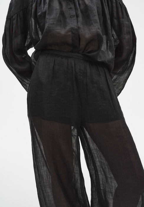Close-up of a sheer black outfit showing a loose blouse tucked into lightweight, semi-transparent trousers with a soft elastic waistband, revealing the silhouette of the legs beneath and emphasizing the airy, textured fabric.