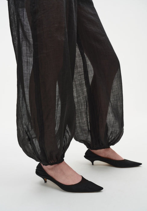 Close-up of sheer black billowy trousers with elasticated cuffs at the ankles, revealing the legs beneath, paired with black pointed low-heel shoes.