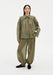 A woman stands facing forward in a relaxed stance against a plain white background, wearing an olive-green sheer blouse with a pointed collar and gathered detailing across the chest, paired with matching lightweight, billowy trousers and dark slip-on shoes.