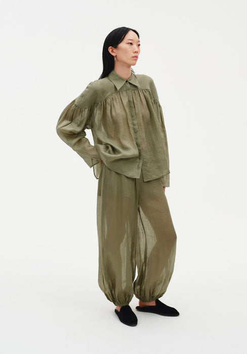 A woman stands in a relaxed three-quarter pose against a plain white background, wearing a loose olive-green sheer blouse with gathered detailing across the chest and matching lightweight trousers with a billowy silhouette, paired with dark slip-on shoes. Her dark shoulder-length hair is worn down, and she looks slightly to the side with one hand resting at her hip.