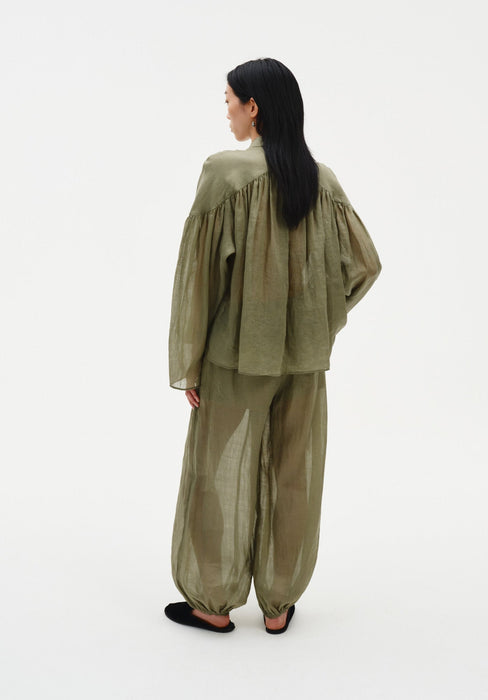 Rear three-quarter view of a woman against a plain white background, wearing a loose olive-green sheer blouse with gathered detailing across the back and matching lightweight trousers with a relaxed, billowy fit.