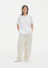 A woman stands facing forward in a relaxed stance against a plain white background, wearing an oversized white T-shirt and loose cream-colored wide-leg trousers with light-colored shoes, her hands in her pockets and posture slightly casual.