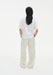 Rear view of a woman standing against a plain white background, wearing a relaxed white short-sleeve top and loose cream-colored trousers. Her dark shoulder-length hair falls straight down her back, and her hands rest in her pockets.