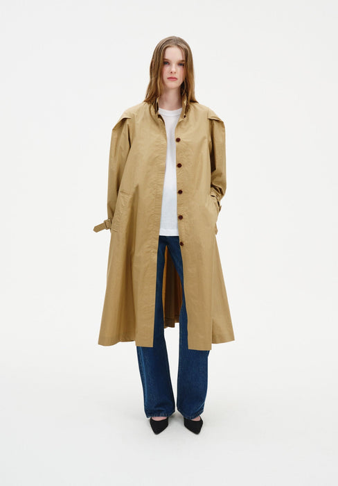 Front view of a model wearing a beige trench-style coat with button front and structured shoulders, layered over a white top and wide-leg blue jeans.