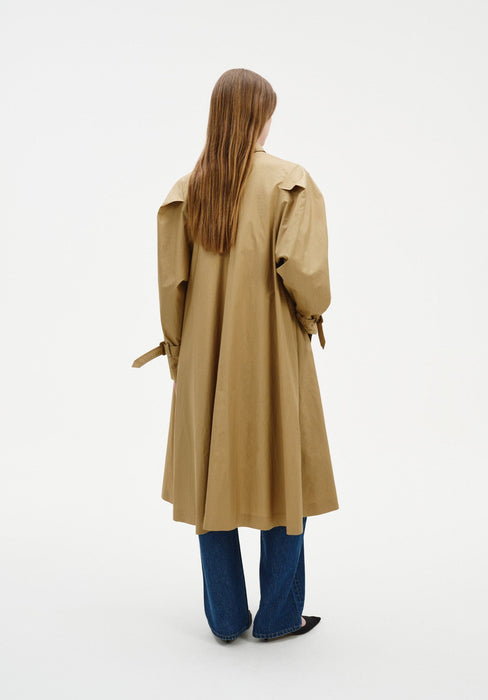Back view of a model wearing a beige trench-style coat with a flared silhouette and cuff straps, paired with wide-leg blue jeans and black pointed shoes.