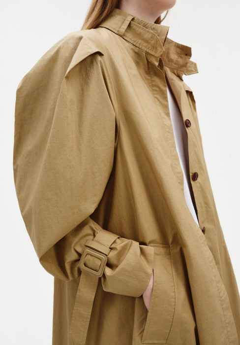 Close-up of a beige trench-style coat highlighting the structured collar, button front, voluminous sleeve, and adjustable cuff strap with buckle, with a hand tucked into the pocket.