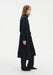 Side profile of a model wearing a long black coat-style dress with a structured collar, relaxed sleeves, and a loose, flowing silhouette, layered over matching wide-leg trousers and paired with pointed black shoes.