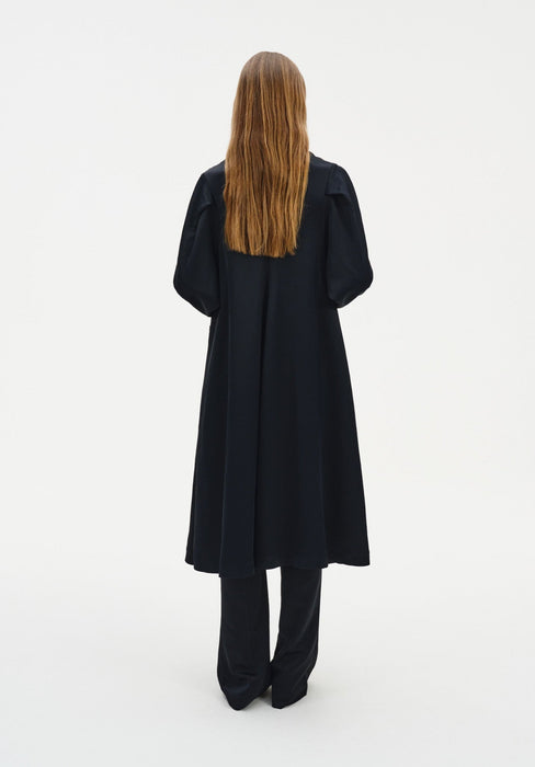 Back view of a model wearing a long black coat-style dress with a clean, flowing silhouette and voluminous sleeves, layered over matching wide-leg trousers against a minimal white background.