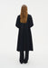 Back view of a model wearing a long black coat-style dress with a clean, flowing silhouette and voluminous sleeves, layered over matching wide-leg trousers against a minimal white background.