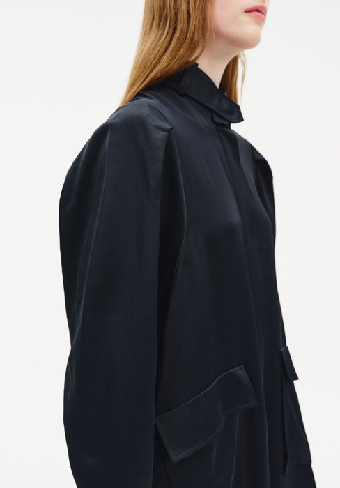 Close-up side view of a black coat-style dress featuring a structured stand collar, subtle sheen fabric, voluminous sleeves, and a large front patch pocket detail.